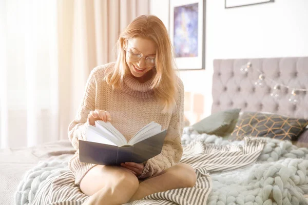 Jeune belle femme en pull chaud livre de lecture sur le lit à la maison, espace pour le texte — Photo