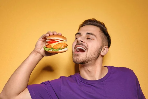 Handsome man eating tasty burger on color background