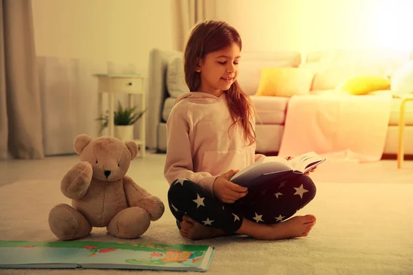 Menina bonito com ursinho sentado no chão e ler livro em casa — Fotografia de Stock