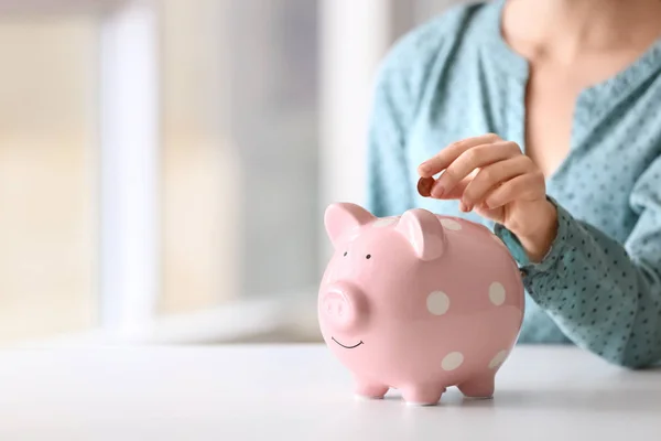 Woman putting coin into piggy bank at table indoors, closeup. Space for text — Stock Photo, Image