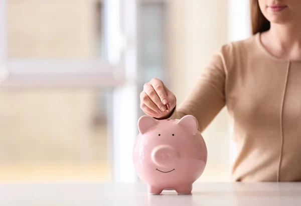 Mujer poniendo moneda en alcancía en la mesa en el interior, primer plano. Espacio para texto — Foto de Stock