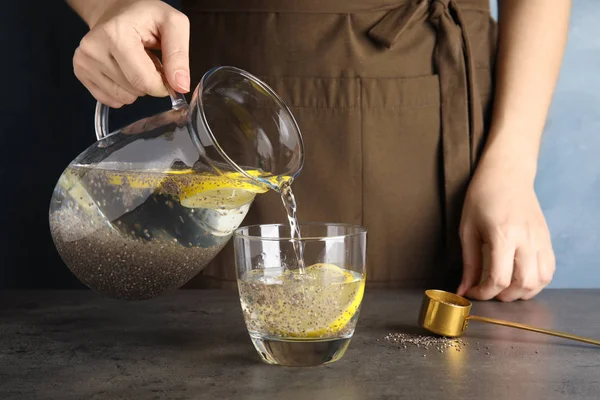 Jovem mulher derramando água com sementes de chia e limão em vidro à mesa, close-up — Fotografia de Stock