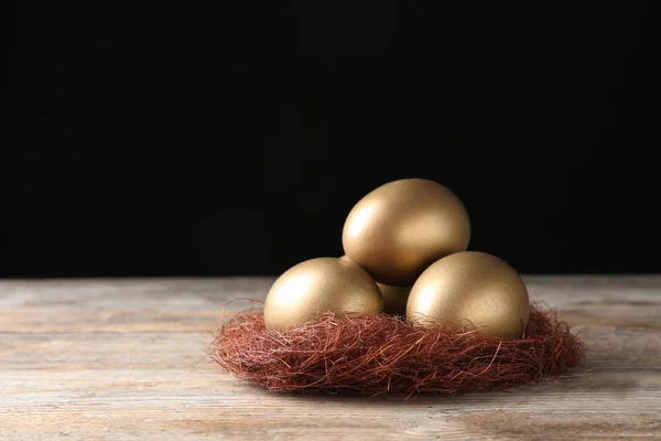 Gouden eieren in nest op houten tafel tegen zwarte achtergrond, ruimte voor tekst — Stockfoto