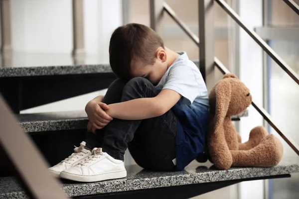 Грустный маленький мальчик с игрушкой сидит на лестнице в помещении — стоковое фото