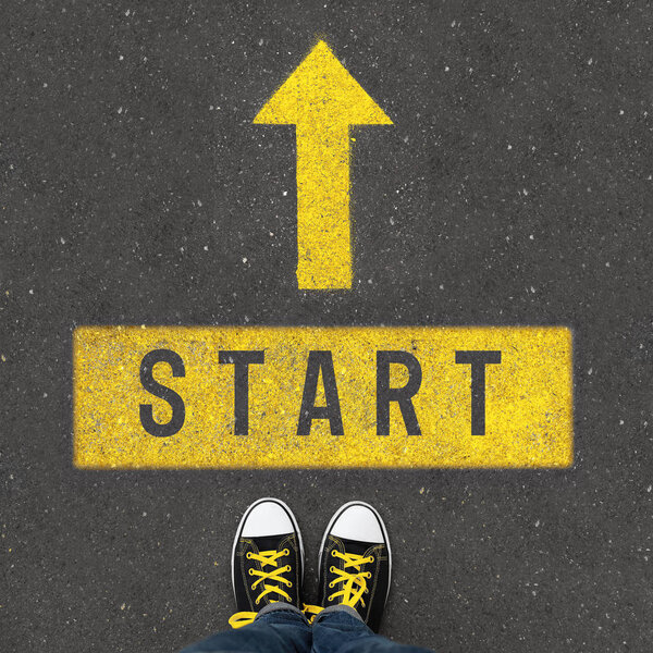 Женщина стоит рядом со стрелкой и слово СТАРТ на асфальте, вид сверху
