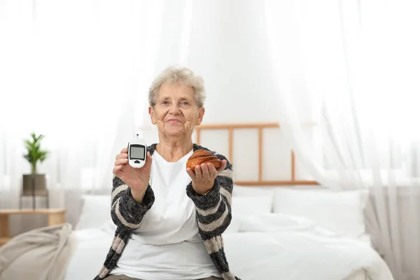 Mujer mayor sosteniendo glucosímetro digital y pastelería en casa. Dieta diabética —  Fotos de Stock