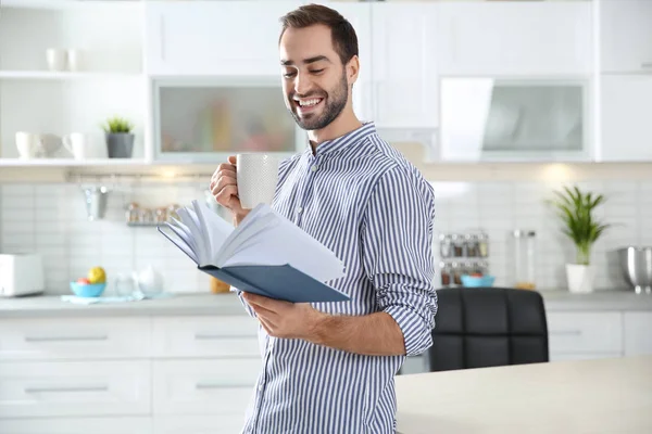 Joven guapo con una taza de libro de lectura de café en casa — Foto de Stock