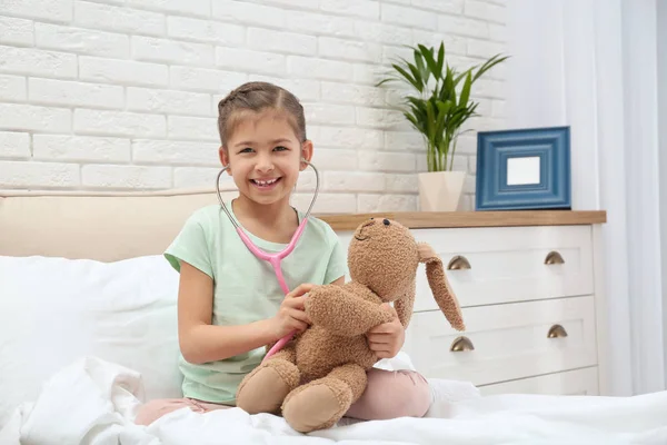 Милый ребенок играет в доктора с мягкой игрушкой на кровати в больничной палате — стоковое фото