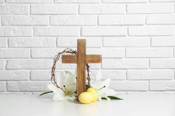 Houten kruis, kroon van doornen, paaseieren en bloesem lelies op tafel tegen bakstenen muur — Stockfoto