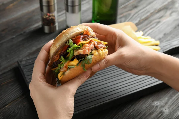 Mulher segurando saboroso hambúrguer com bacon na mesa, close-up — Fotografia de Stock