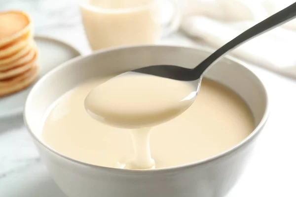 Spoon of pouring condensed milk over bowl on table, closeup. Dairy products — Stock Photo, Image