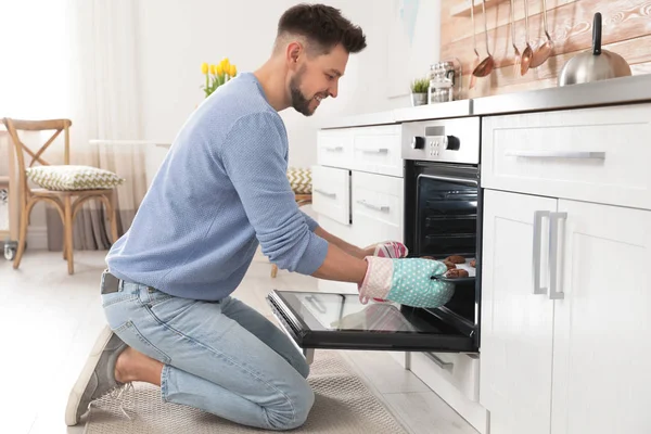 Handsome man taking out tray of baked cookies from oven in kitchen — Stock Photo, Image