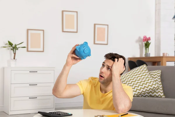 Sad man looking for money in piggy bank at table indoors