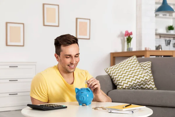 Χαρούμενος άνθρωπος βάζοντας κέρματα σε κουμπαράς στο τραπέζι στο σαλόνι. Εξοικονόμηση χρημάτων — Φωτογραφία Αρχείου