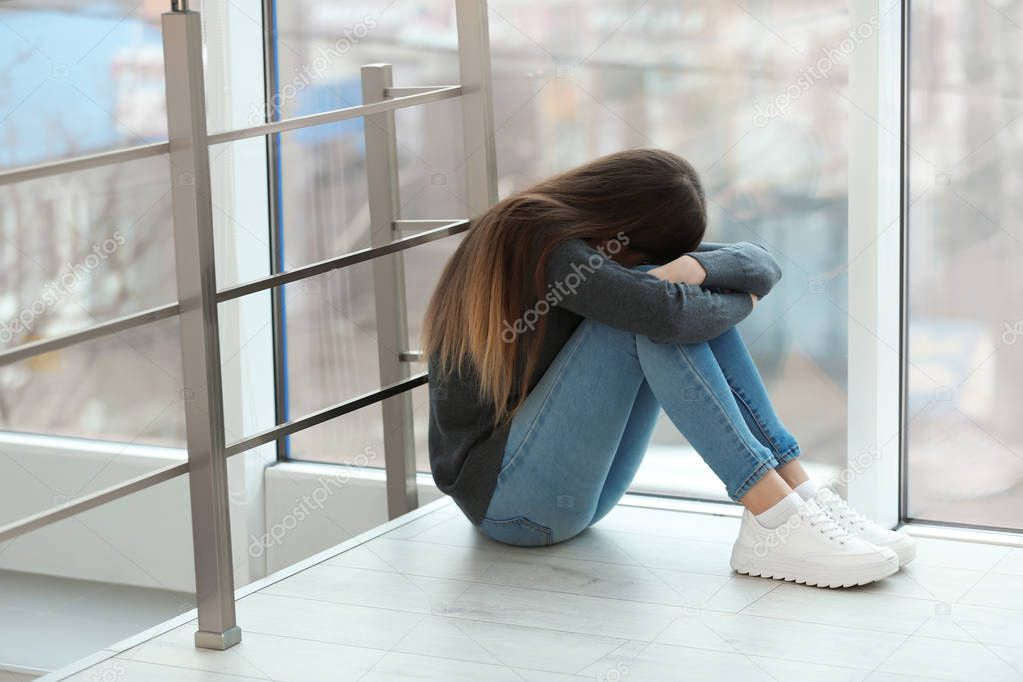Upset teenage girl sitting at window indoors