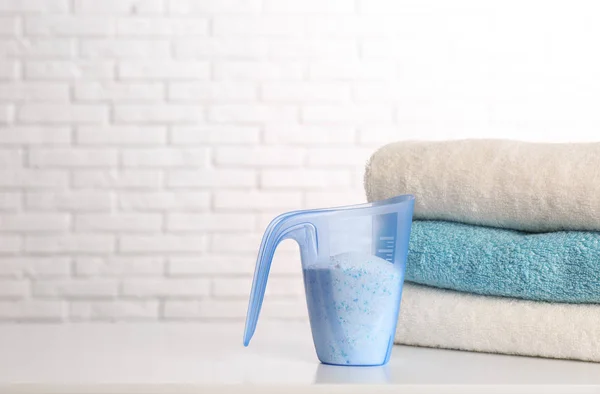 Measuring cup with washing powder and clean towels on table against brick wall, space for text. Laundry day — Stock Photo, Image