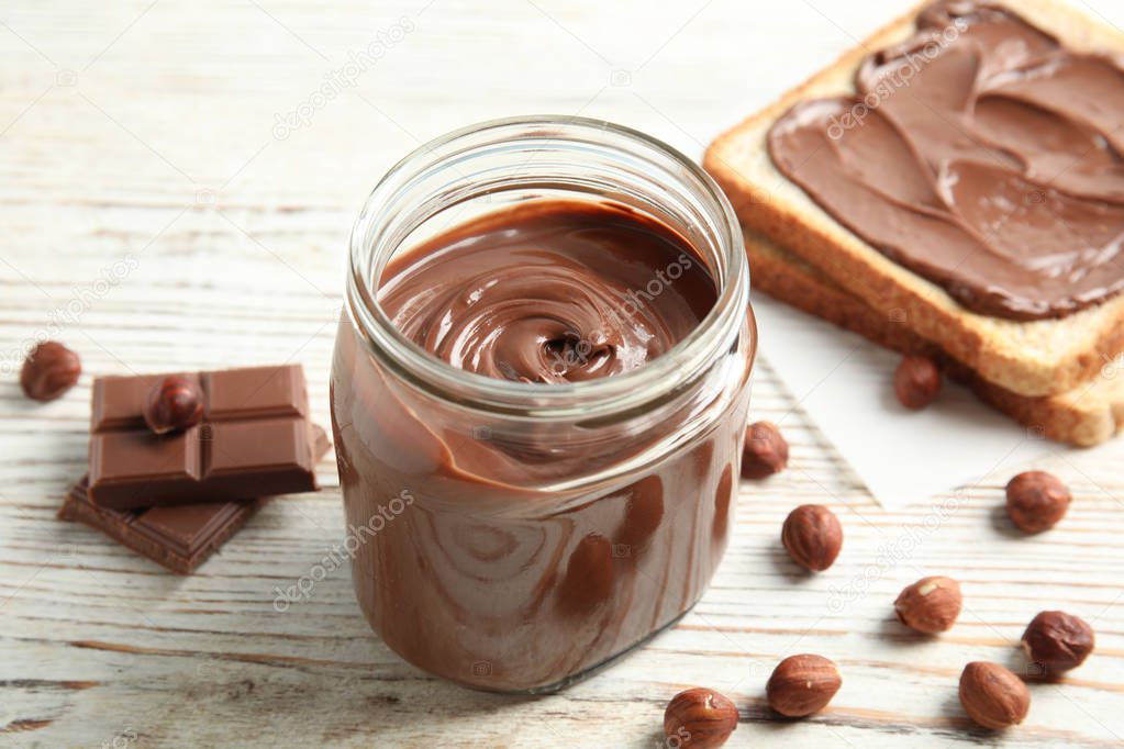 Glass jar with tasty chocolate cream, bread and hazelnuts on wooden table