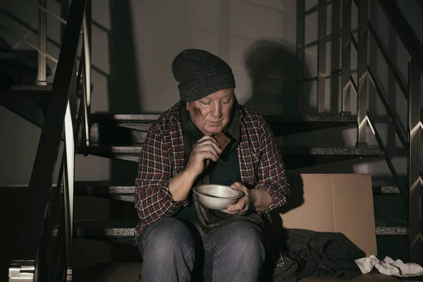Homem sênior pobre com tigela e pão nas escadas dentro de casa — Fotografia de Stock