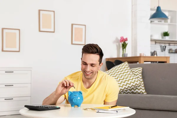 Hombre feliz poniendo moneda en alcancía en la mesa en la sala de estar. Ahorro de dinero —  Fotos de Stock