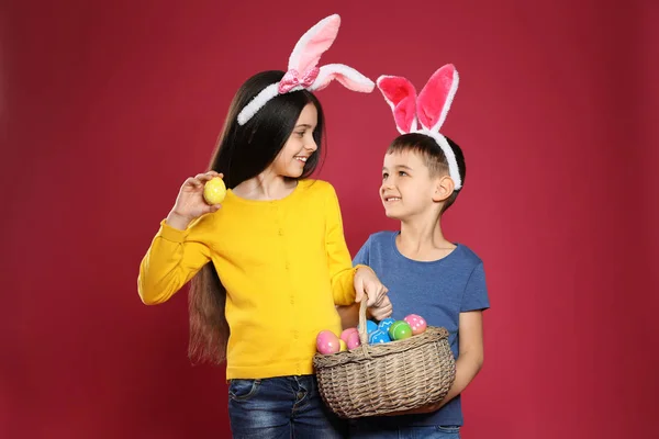 Cute children in bunny ears headbands holding basket with Easter eggs on color background — Stock Photo, Image