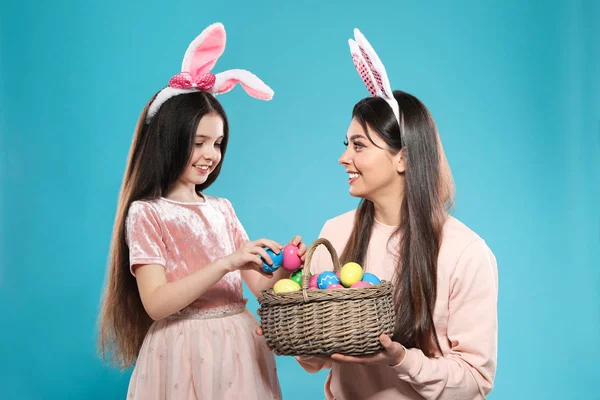 Mother and daughter in bunny ears headbands with basket of Easter eggs on color background — Stock Photo, Image