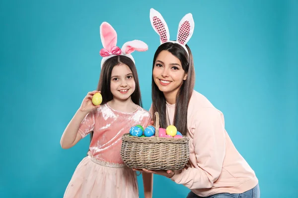 Mother and daughter in bunny ears headbands with basket of Easter eggs on color background — Stock Photo, Image