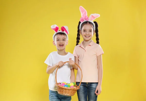 Niedlichen Kindern in Hasenohren Stirnbänder halten Korb mit Ostereiern auf farbigem Hintergrund — Stockfoto