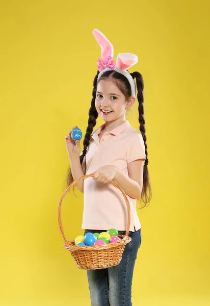 Little girl in bunny ears headband holding basket with Easter eggs on color background — Stock Photo, Image