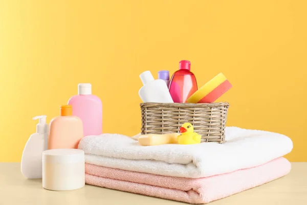 Wicker basket with baby cosmetic products, toy and towels on table against color background