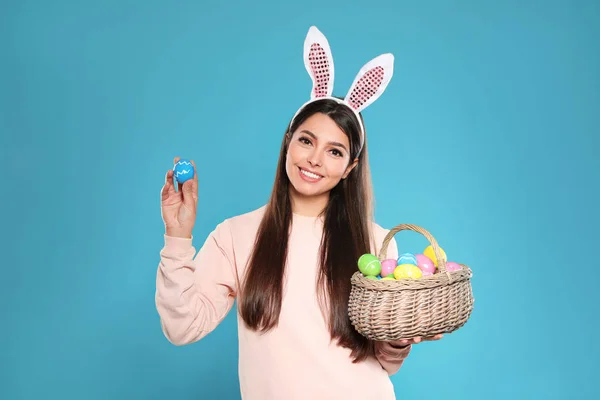 Beautiful woman in bunny ears headband holding basket with Easter eggs on color background — Stock Photo, Image