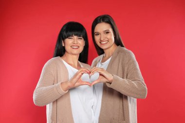 Genç kız ve olgun annesi, elleri renk arka planda kalp yapıyor.