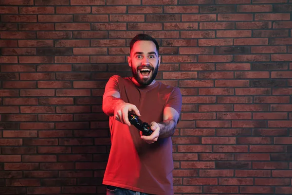 Hombre emocional jugando videojuegos con controlador cerca de la pared de ladrillo —  Fotos de Stock