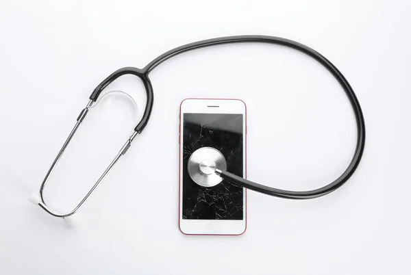 Nowoczesny smartfon ze złamanym wyświetlaczem i stetoskop na białym tle, widok z góry. Naprawa urządzeń — Zdjęcie stockowe