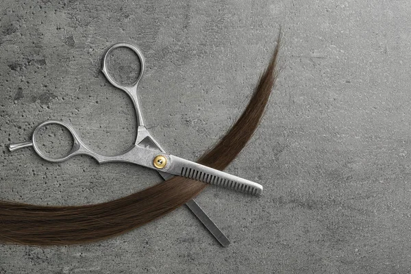 Composición plana con hebra de pelo castaño y tijeras de adelgazamiento sobre fondo gris. Servicio de peluquería —  Fotos de Stock