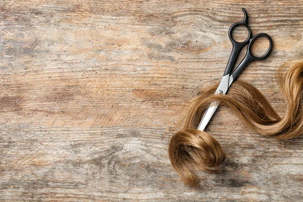 Flat lay composition with strand of light brown hair, scissors and space for text on wooden background. Hairdresser service