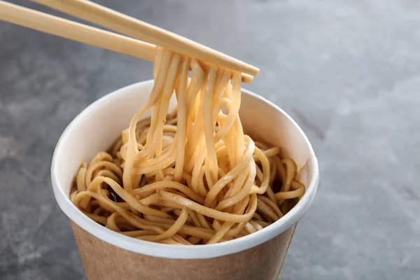 Tazza di carta di pasta istantanea con salsa e bacchette su sfondo grigio, primo piano. Spazio per testo — Foto Stock