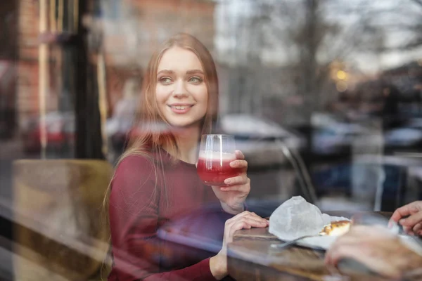 Όμορφη νεαρή γυναίκα με κοκτέιλ και κέικ στο τραπέζι στο καφέ, θέα από έξω από το παράθυρο — Φωτογραφία Αρχείου
