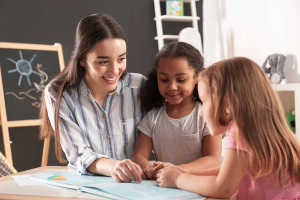 Crianças pequenas bonitos com professor de berçário livro de leitura à mesa no jardim de infância. Actividades internas — Fotografia de Stock