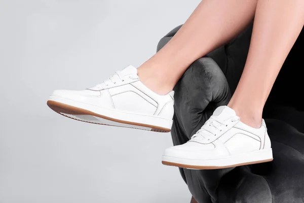 Woman with stylish sport shoes sitting in armchair on light background