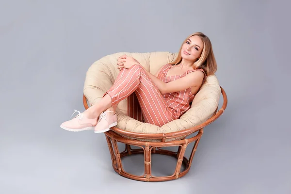 Νεαρή κομψή γυναίκα με μοντέρνα παπούτσια κάθεται σε καρέκλα papasan σε γκρι φόντο — Φωτογραφία Αρχείου