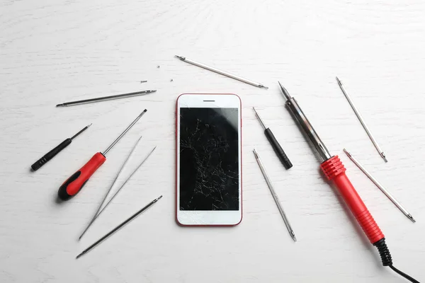 Mobile phone with broken display and repair tools on wooden table, flat lay