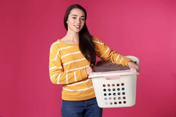 Happy young woman holding basket with laundry on color background