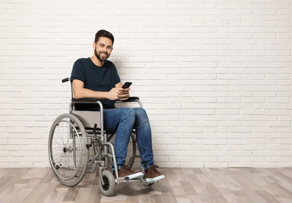 Jongeman met smartphone in rolstoel in de buurt van bakstenen muur binnen. Ruimte voor tekst — Stockfoto
