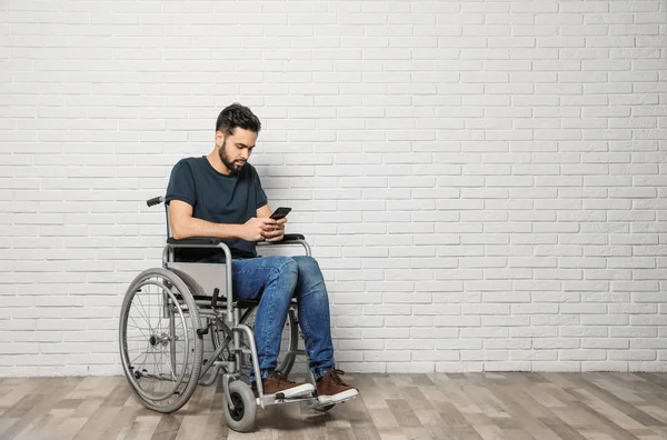 Młody człowiek ze smartfonem na wózku inwalidzkim przy ścianie z cegły. Miejsce na tekst — Zdjęcie stockowe