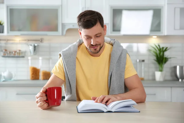 Hombre guapo con taza de libro de lectura de café en la mesa en la cocina — Foto de Stock
