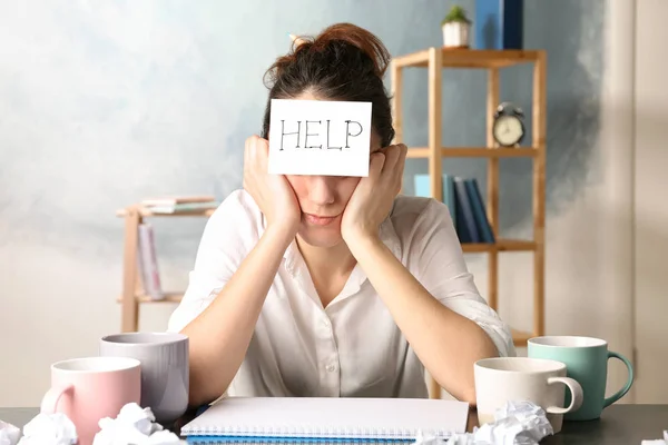 Young woman with note HELP on forehead at workplace — 스톡 사진