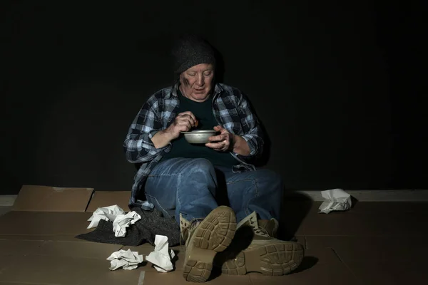 Poor senior man with bread and bowl on floor near dark wall — ストック写真