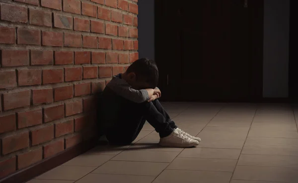 Triste menino sentado no chão perto da parede de tijolo dentro de casa — Fotografia de Stock