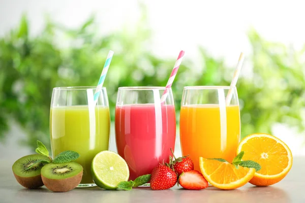 Glasses of different juices with straws and fresh fruits on table against blurred background — Stock Photo, Image