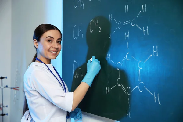 Female scientist writing chemical formula on chalkboard indoors — Stock Photo, Image